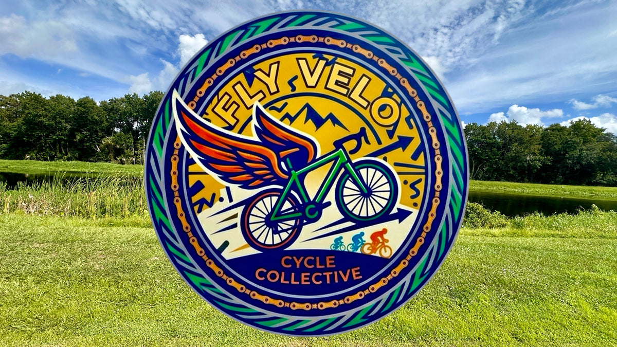 A large circular Fly Velo Cycle Collective logo sign displayed outdoors on a green grassy field under a blue sky with clouds.